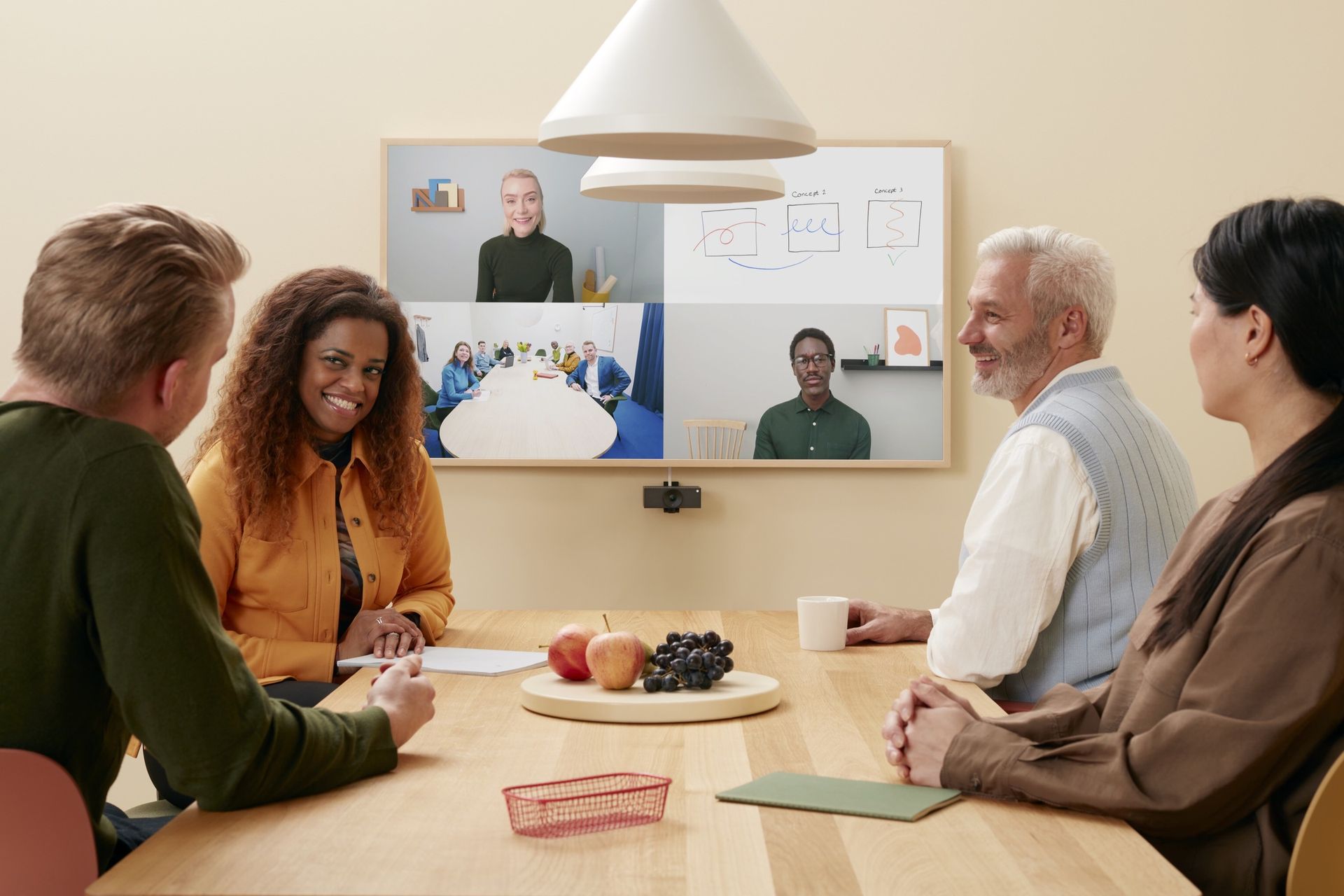 A meeting room in use: five people around a table, with remote participants on a wall display and a camera framing the table.