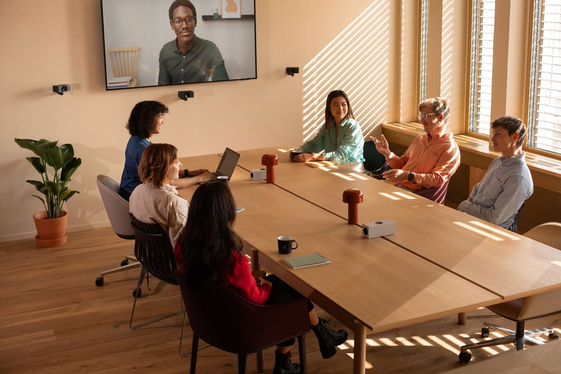 A meeting room mid-call: five people around a table with two remote participants on the wall display above a video bar, and a camera framing the room.